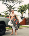 Man holding a surfboard with a beach towel and palm trees in the background