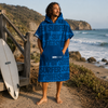 A man stands barefoot on a beach path wearing a blue surf-themed hooded towel poncho. The garment has a large front pocket and the word 'SURFER' printed repeatedly in varying shades of blue.