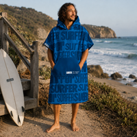 A man stands barefoot on a beach path wearing a blue surf-themed hooded towel poncho. The garment has a large front pocket and the word 'SURFER' printed repeatedly in varying shades of blue.